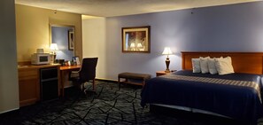 A hotel room with a bed, desk, microwave, chair, and a small table with a lamp.