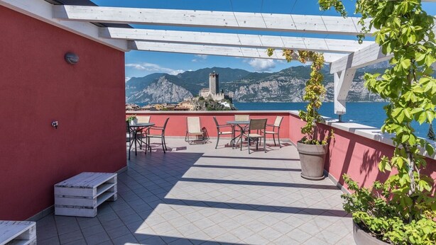 A terrace with outdoor seating, a view of the sea, and a castle in the background. - Residence "La Pergola" central area, garden, swimming pool, parking (Malcesine)