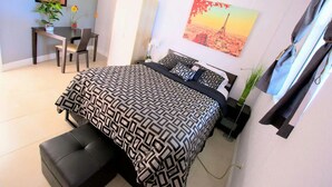 Una camera da letto con una coperta a motivi, una scrivania con una sedia, una pianta in vaso e un quadro raffigurante un paesaggio urbano.