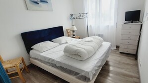 A neatly made bed with white linens, a wooden chair, a bedside table with a lamp, and a TV on a dresser.