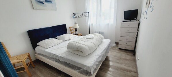 A neatly made bed with white linens, a wooden chair, a bedside table with a lamp, and a TV on a dresser.