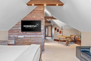 A loft-style bedroom with exposed wooden beams, a mounted TV, a desk, and a sofa.