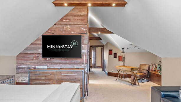 Ein Schlafzimmer im Loft-Stil mit freiliegenden Holzbalken, einem Wand-Fernseher, einem Schreibtisch und einem Sofa.