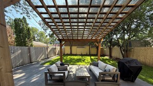 Eine überdachte Terrasse mit Holzmöbeln, einem Grill und Blick auf einen Garten.