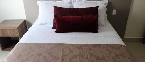 A neatly made bed with white bedding and a single dark red pillow, a wooden nightstand to the left, and a patterned bedspread.
