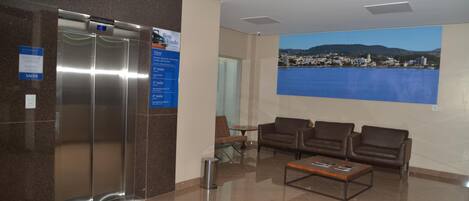 A lobby with a reception desk, a digital clock, a large window with a cityscape view, and a seating area with sofas.