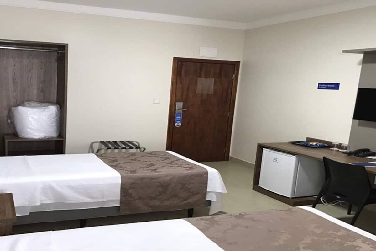 Habitación de hotel con dos camas individuales, un escritorio con computadora, televisión y un refrigerador pequeño.