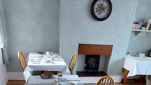 Un coin repas avec une longue table dressée pour deux personnes, une cheminée et une horloge au mur.