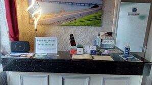 Un comptoir d’accueil avec un panneau indiquant les allergies et intolérances alimentaires, un mur décoratif orné d’un tableau représentant un paysage, et un présentoir avec divers articles.