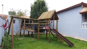 A wooden play structure with a slide and climbing net, surrounded by a grassy yard and a blue building with a tiled roof. - Sítio em Araçoiaba da Serra (Araçoiaba da Serra)