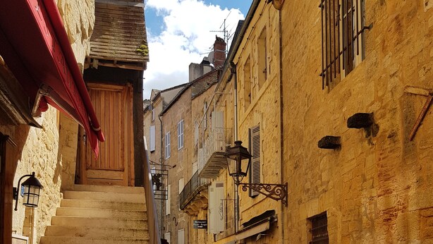 Une rue étroite bordée d’immeubles en pierre, avec des tables installées à l’extérieur et un escalier menant à un niveau supérieur.
