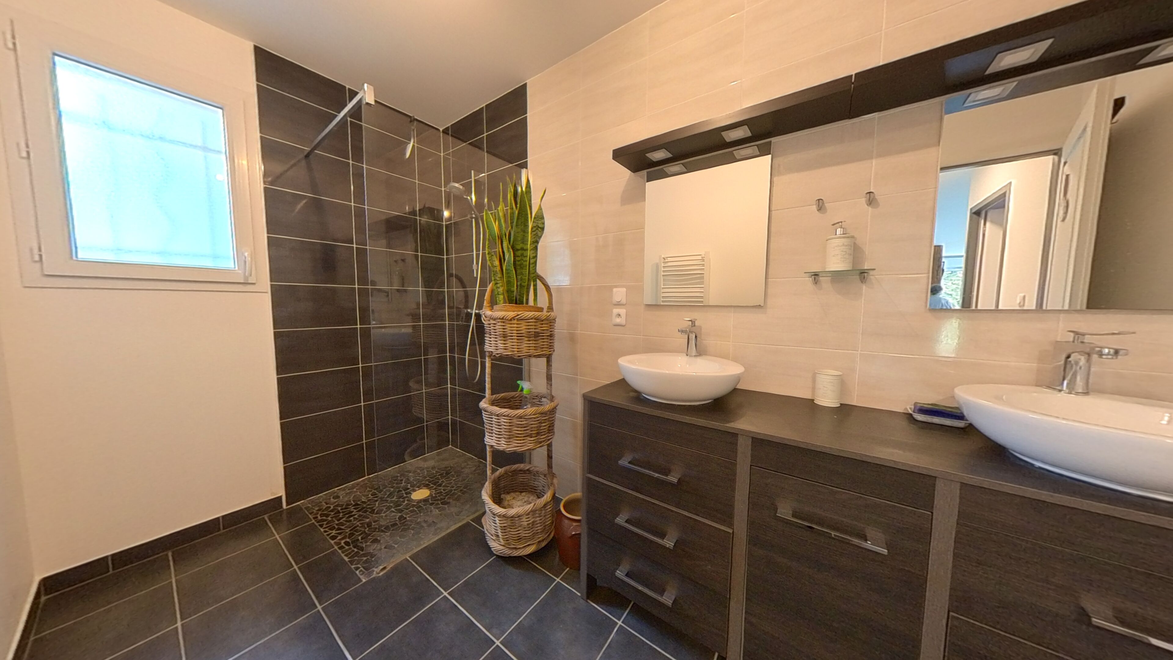 A modern bathroom with a walk-in shower, two sinks, dark tiled walls, and a large mirror.