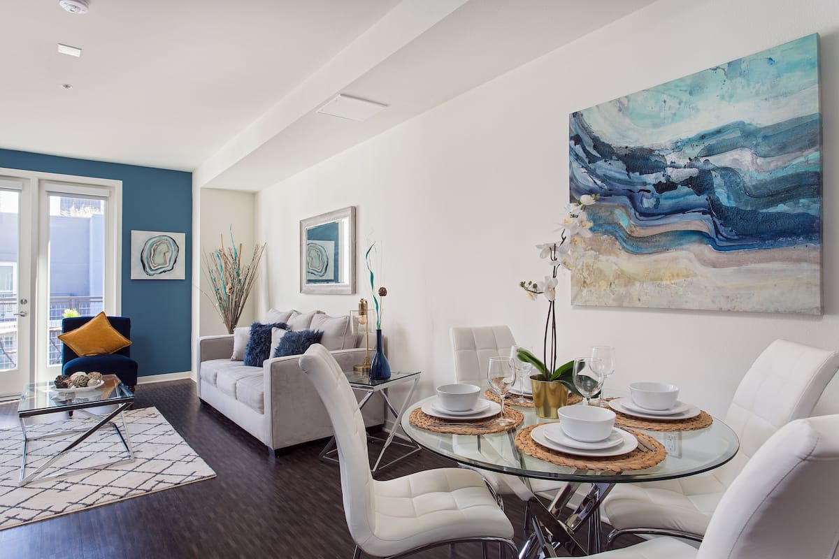 Ein modernes Wohnzimmer mit einem Esstisch aus Glas, gedeckt für vier Personen, weißen Sofas und einem großen abstrakten Gemälde an der Wand.