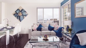 A modern living room with a glass coffee table, a sofa with floral cushions, a dining area with a glass table, and a large window with a city view. - New Downtown LA Luxury Jr. Penthouse (Los Angeles)