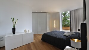 Een moderne slaapkamer met een groot bed, een nachtkastje met lamp, een dressoir en uitzicht op groen.