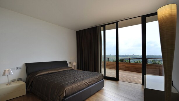 Un dormitorio moderno con una cama grande, una mesita de noche, una lámpara y vistas a un balcón con barandilla.