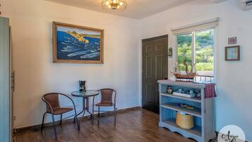 A room with a wooden floor, two wicker chairs, a small table, a painting of a ship, a bookshelf with nautical items, and a window with a view of greenery.
