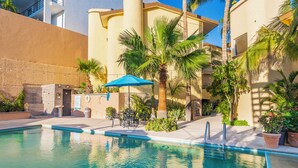A swimming pool with a blue umbrella, palm trees, and a building with a beige facade. - Antigua de Cabo (Cabo San Lucas)