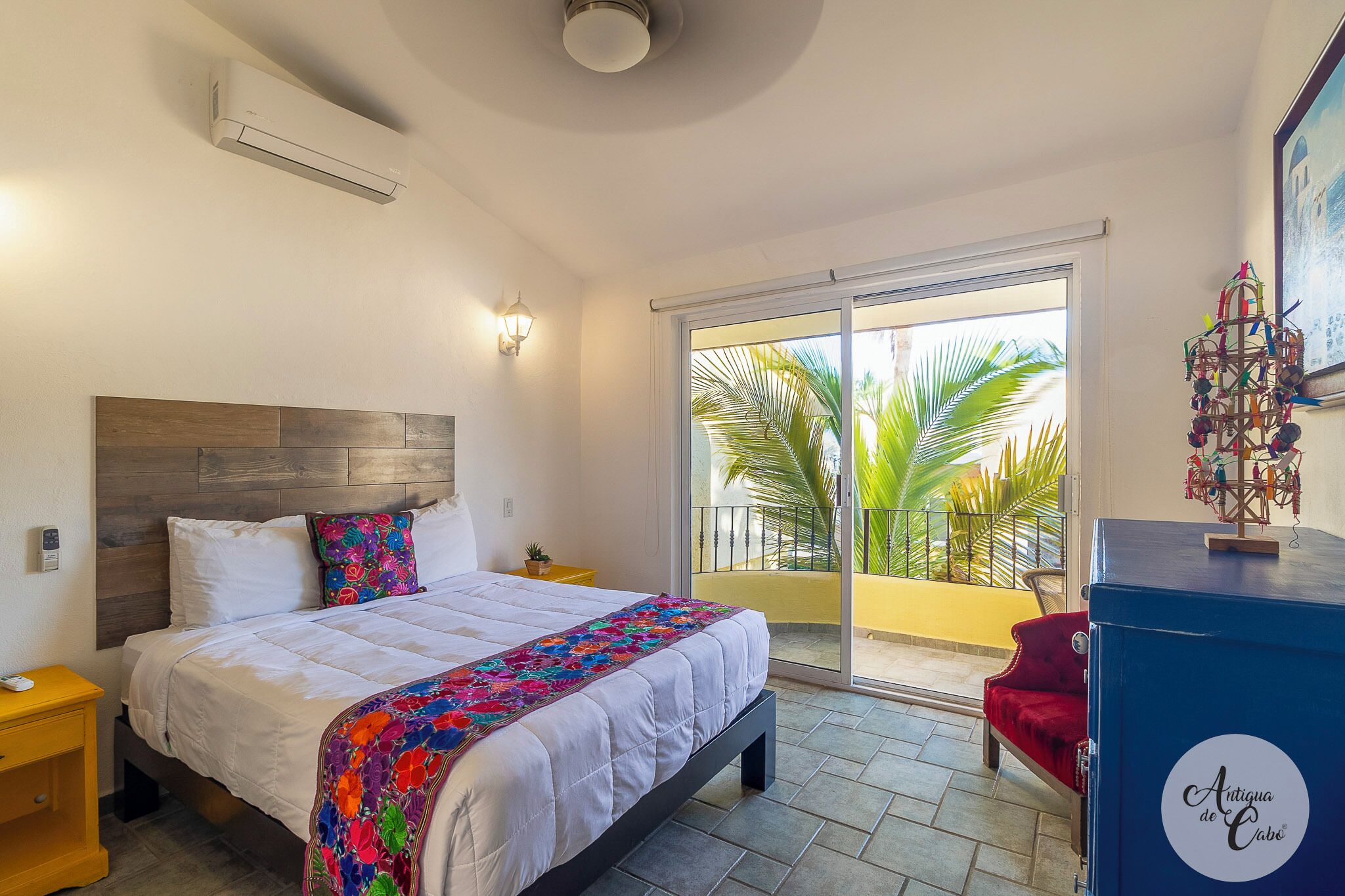 Een slaapkamer met een bed, een nachtkastje, een rode stoel, een balkon met uitzicht op palmbomen, en een decoratief object aan de muur.