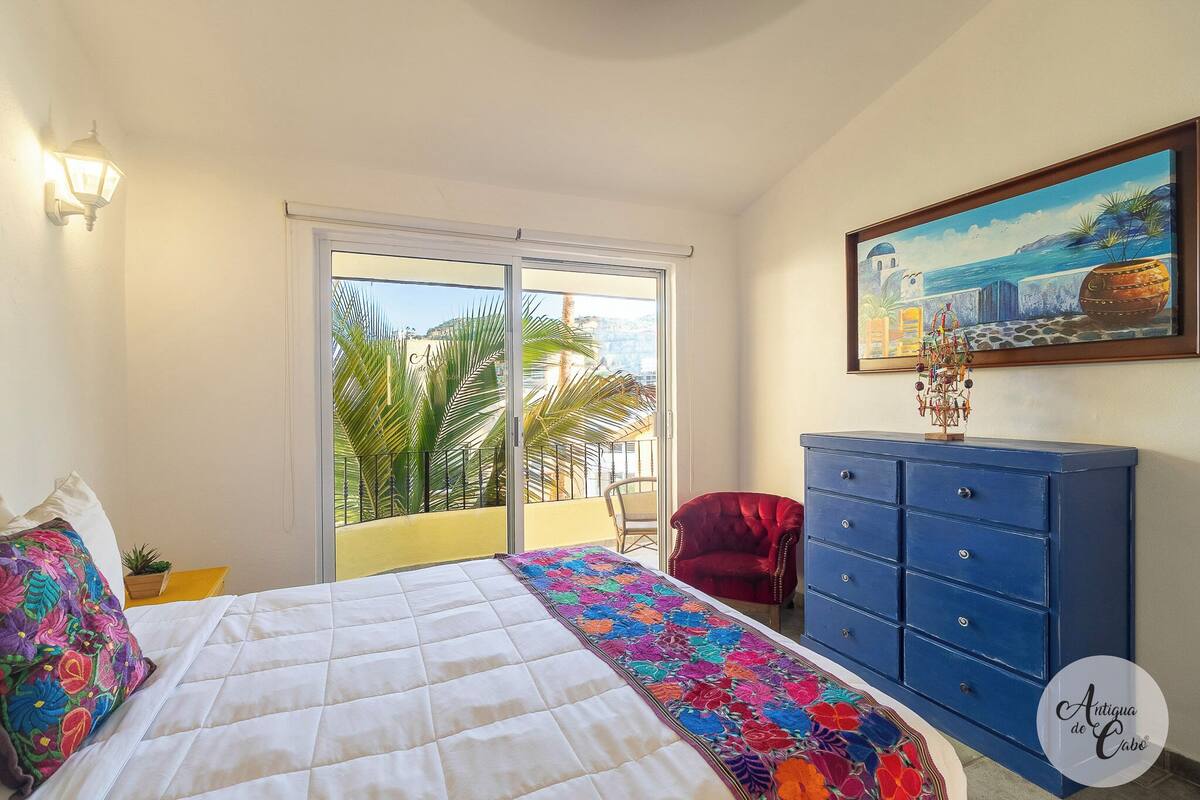 Un dormitorio con cama, cómoda azul, silla roja, un cuadro y balcón con vista a palmeras y al mar.