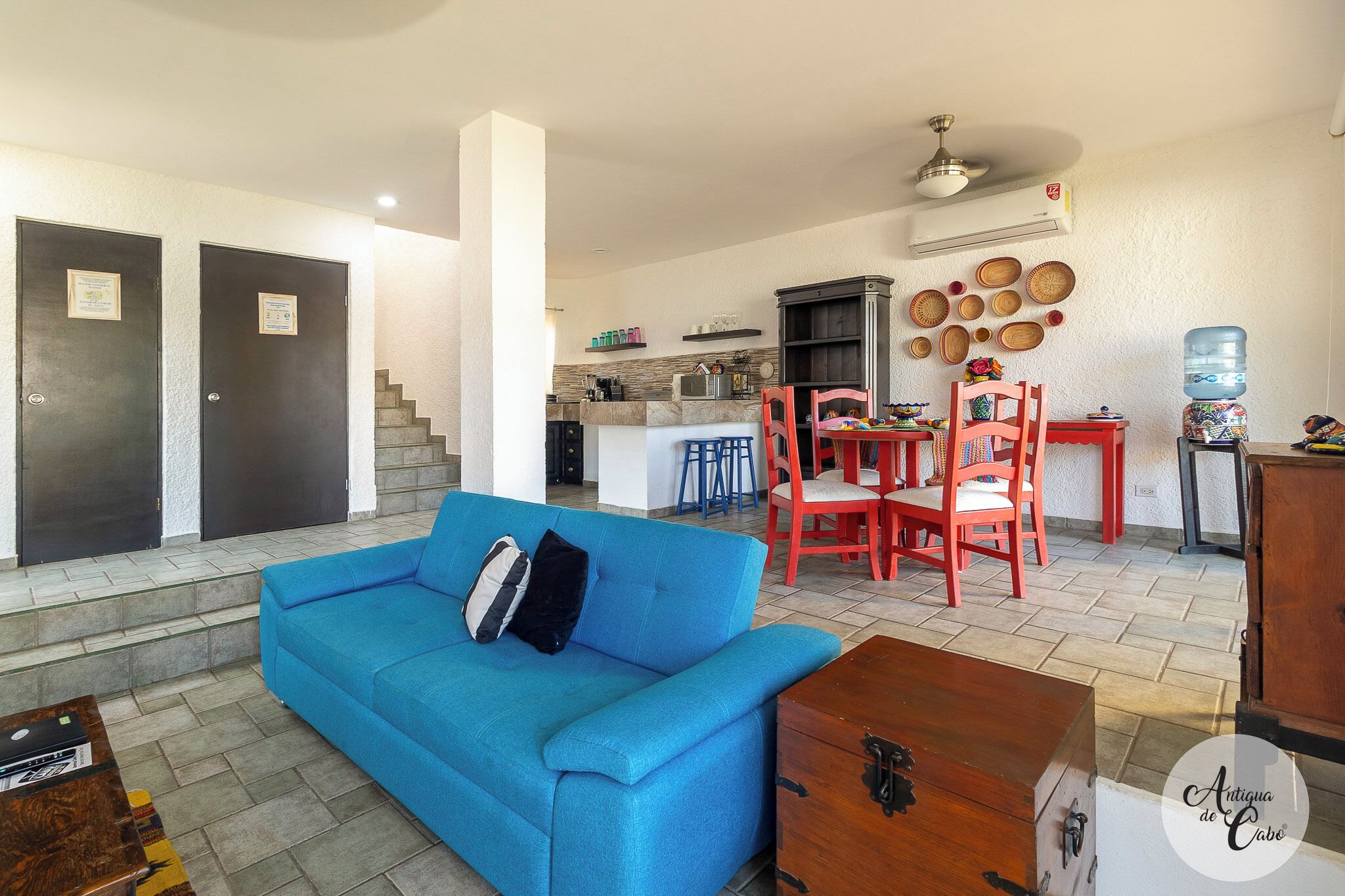 A living room with a blue sofa, a wooden coffee table, and a red dining table with chairs. There is a kitchen area in the background with a water dispenser and a refrigerator.