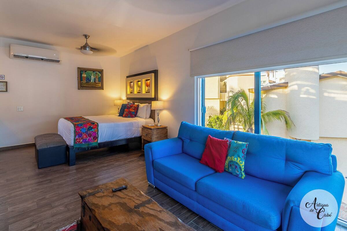 Una sala de estar moderna con un sofá azul, una cama con ropa de cama colorida, una mesa de centro de madera y un ventanal con vistas a palmeras.