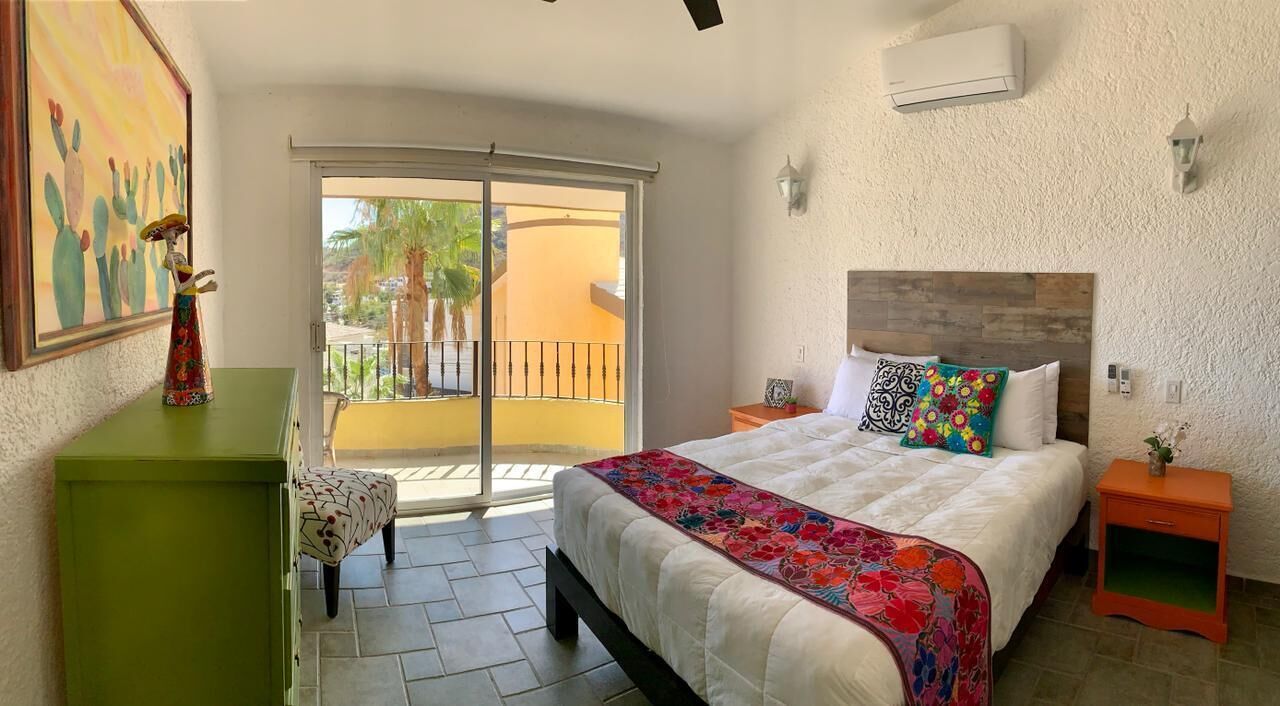 A bedroom with a bed, a nightstand, a green dresser, a chair, and a balcony with a view of palm trees and a building.