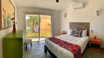 A bedroom with a bed, a nightstand, a green dresser, a chair, and a balcony with a view of palm trees and a building.