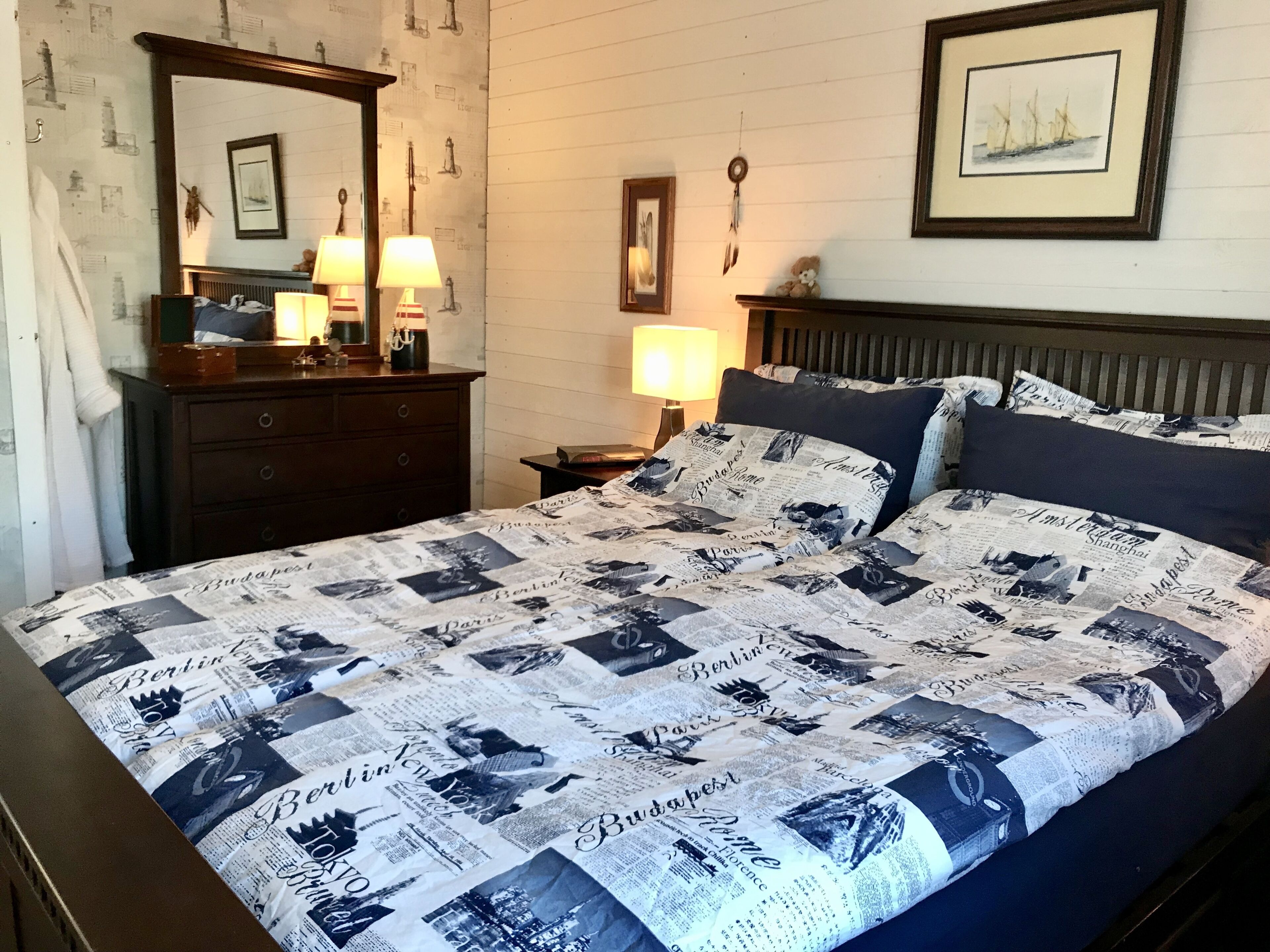 Habitación de hotel con una cama que presenta un edredón con motivos de paisaje urbano, un aparador, una mesita de noche con una lámpara y un cuadro en la pared.