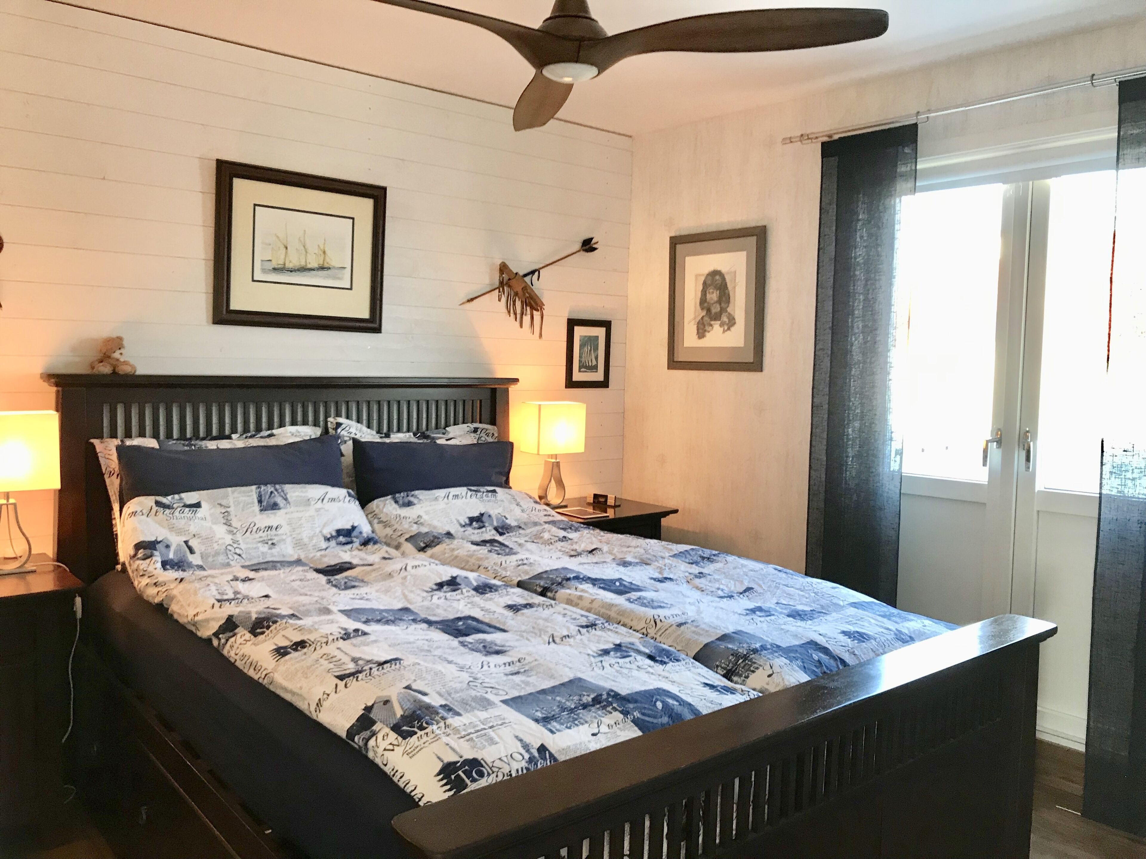 A bedroom with a bed, two bedside tables, a ceiling fan, and framed pictures on the walls.