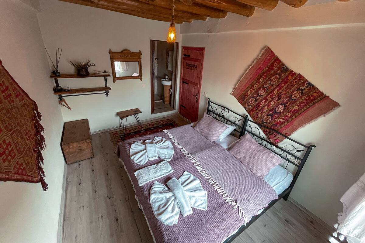 A bedroom with a wooden ceiling, a bed with a patterned bedspread, a red door, and a large patterned rug.