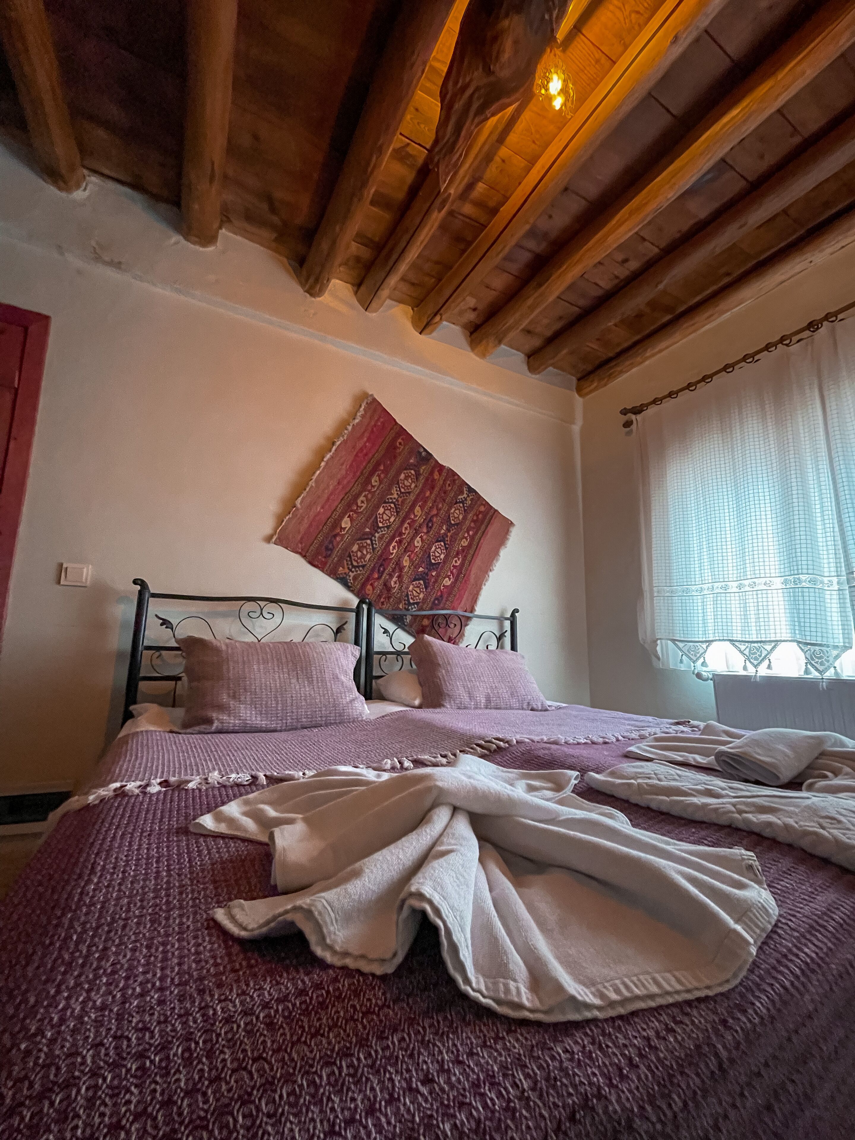 A bedroom with a wooden ceiling, a bed with a patterned headboard, and a window with a sheer curtain.