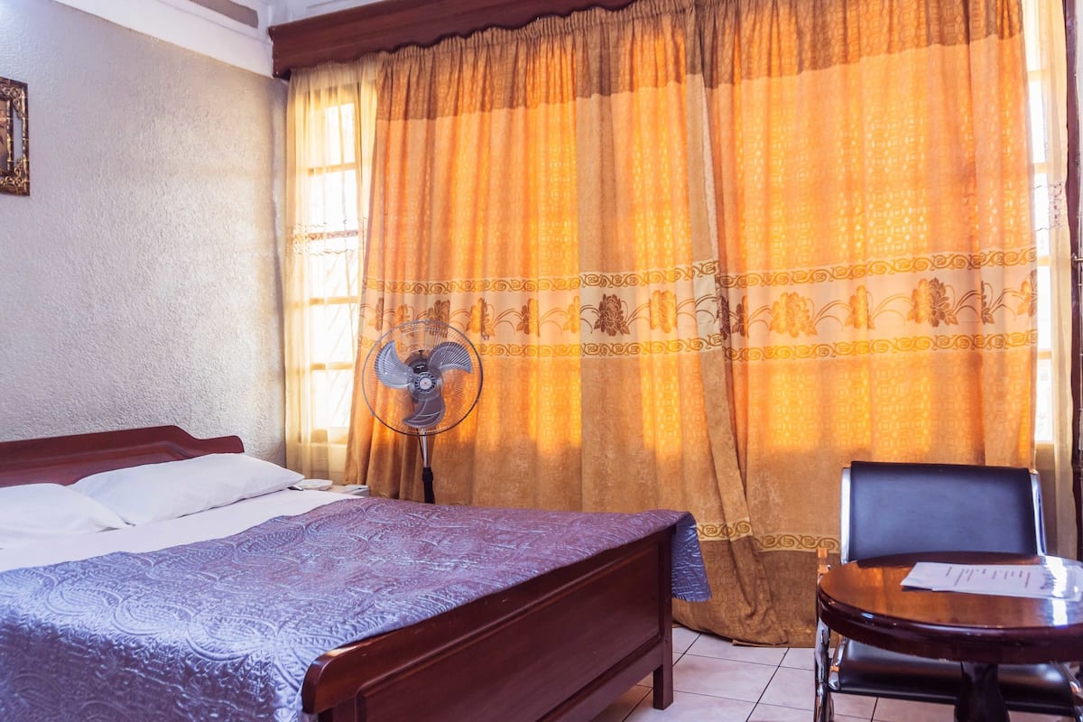 A bedroom with a bed, a chair, a standing fan, and a window with curtains.
