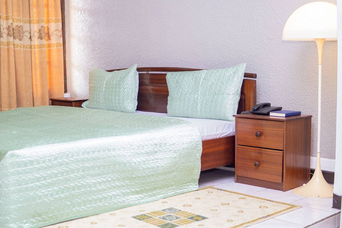 A neatly made bed with a green bedspread, a wooden nightstand with a lamp, and a patterned rug on the floor.