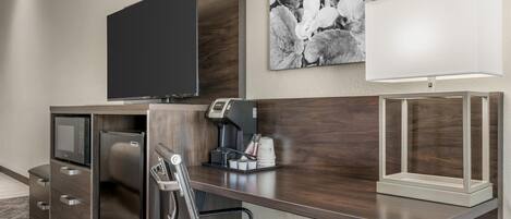 Et hotelværelse med et fladskærms-tv, kaffemaskine, mikroovn, køleskab, en stol og en lampe.
