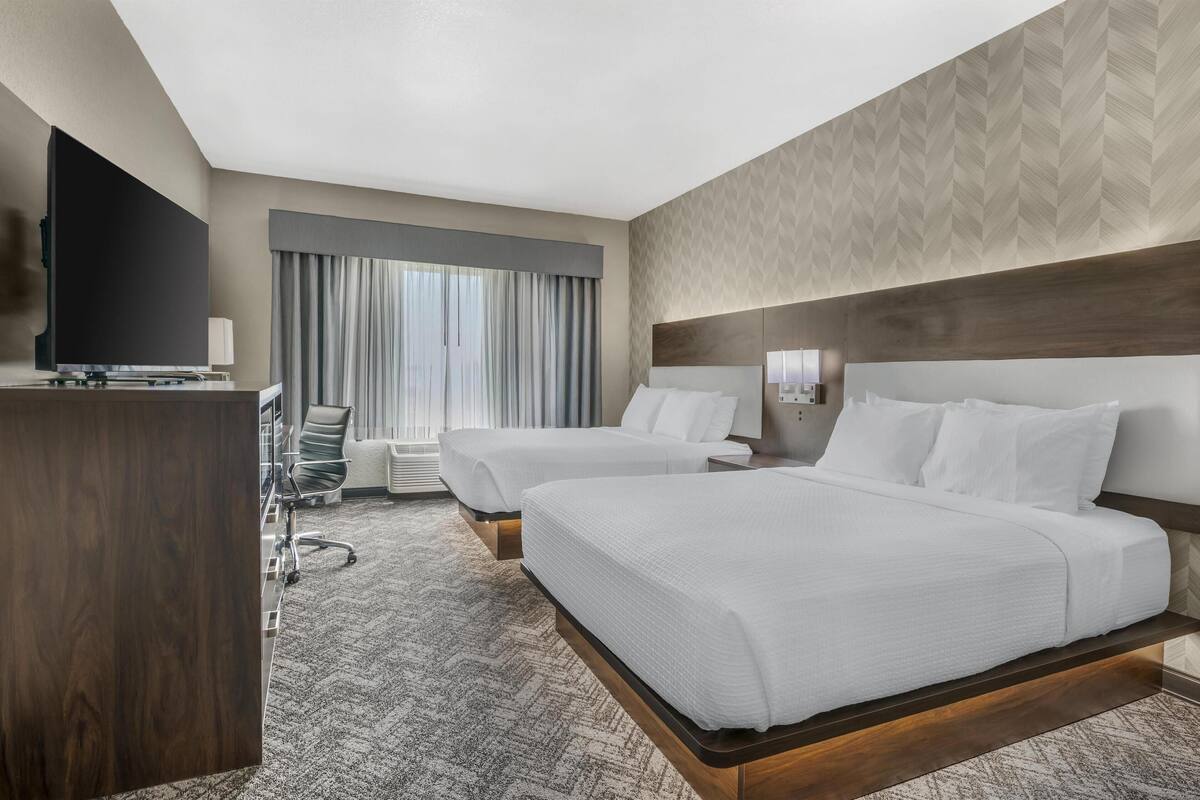 Habitació d'hotel amb dos llits, un escriptori, una cadira, un televisor i una finestra amb cortines.
