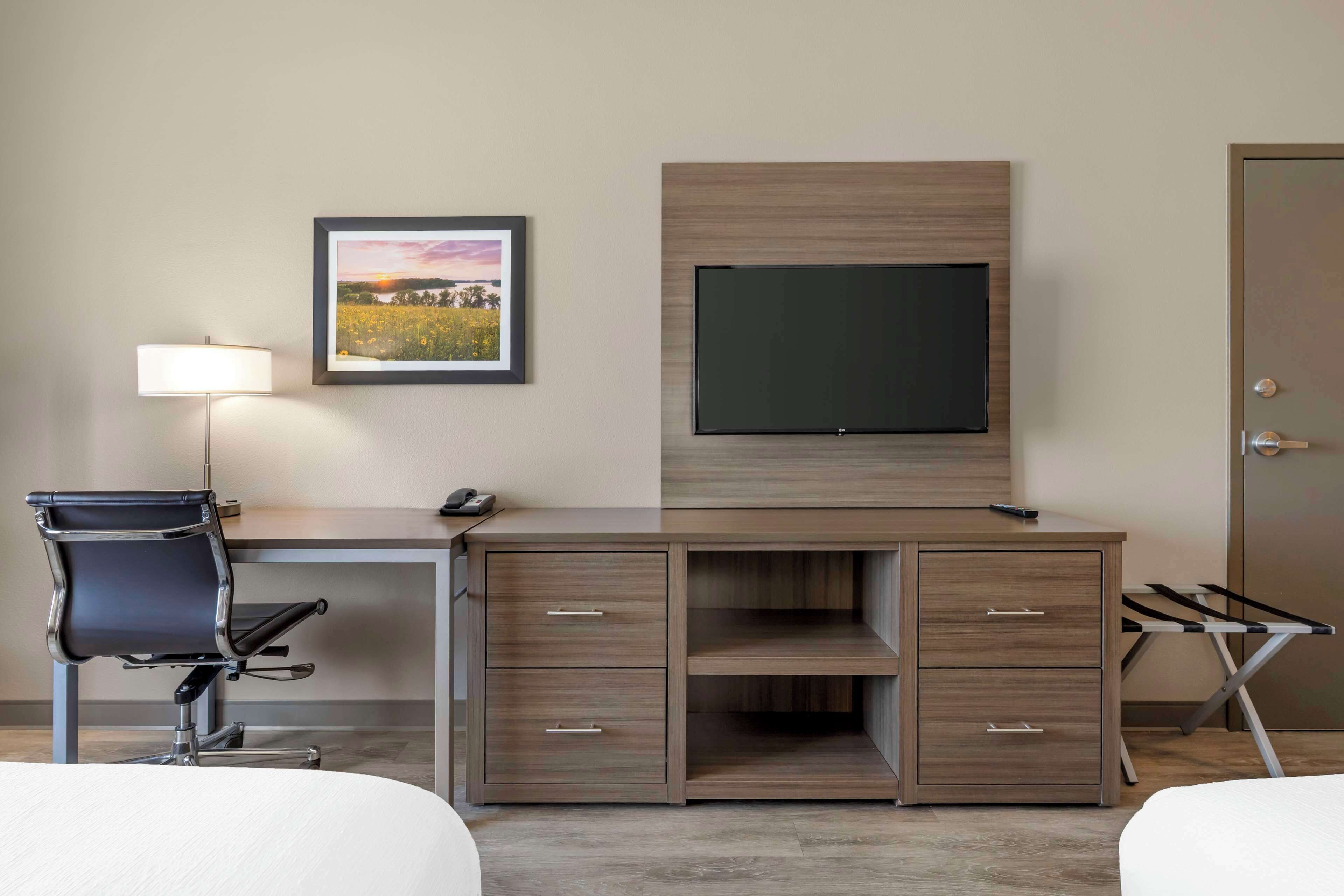 A hotel room with a desk, a chair, a TV mounted on the wall, a framed picture, and a door.