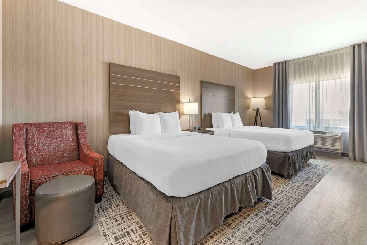 A hotel room with two beds, a red armchair, a small round table, and a lamp.