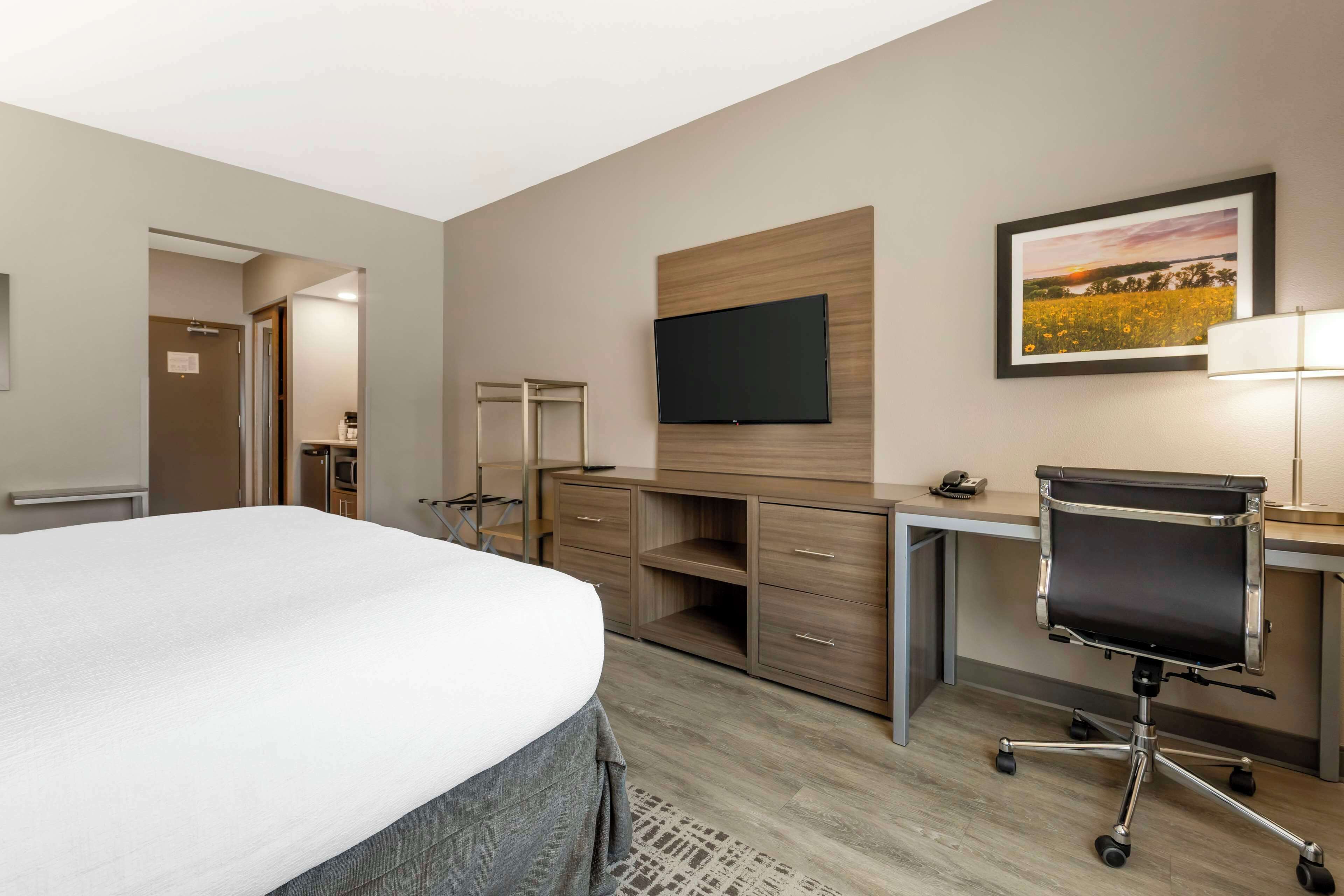 A hotel room with a bed, a desk with a chair, a flat-screen TV, a wooden cabinet, and a framed picture on the wall.