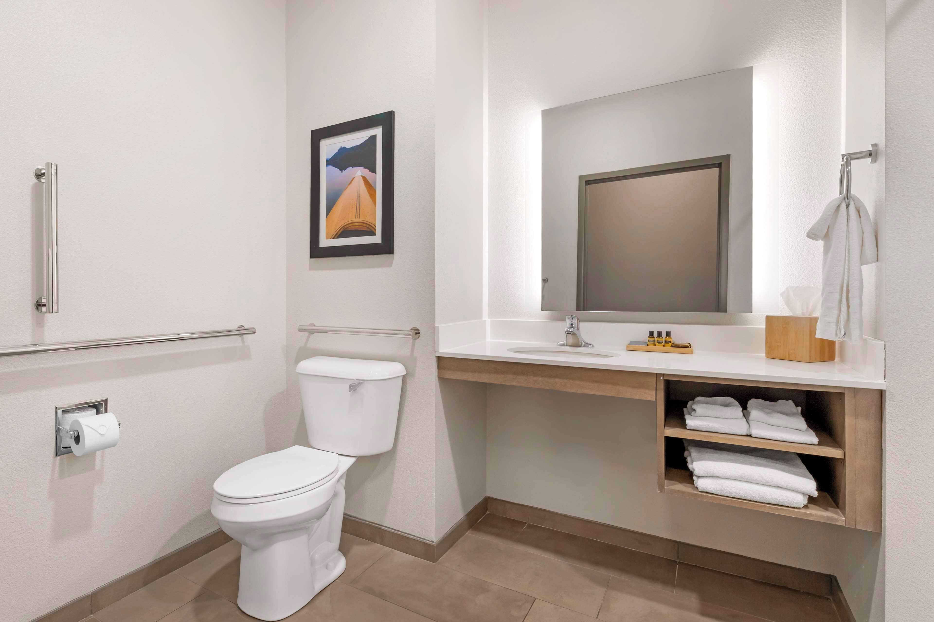 Une salle de bain moderne équipée de toilettes, d’une barre d’appui, d’un lavabo avec un miroir et d’une étagère avec des serviettes.