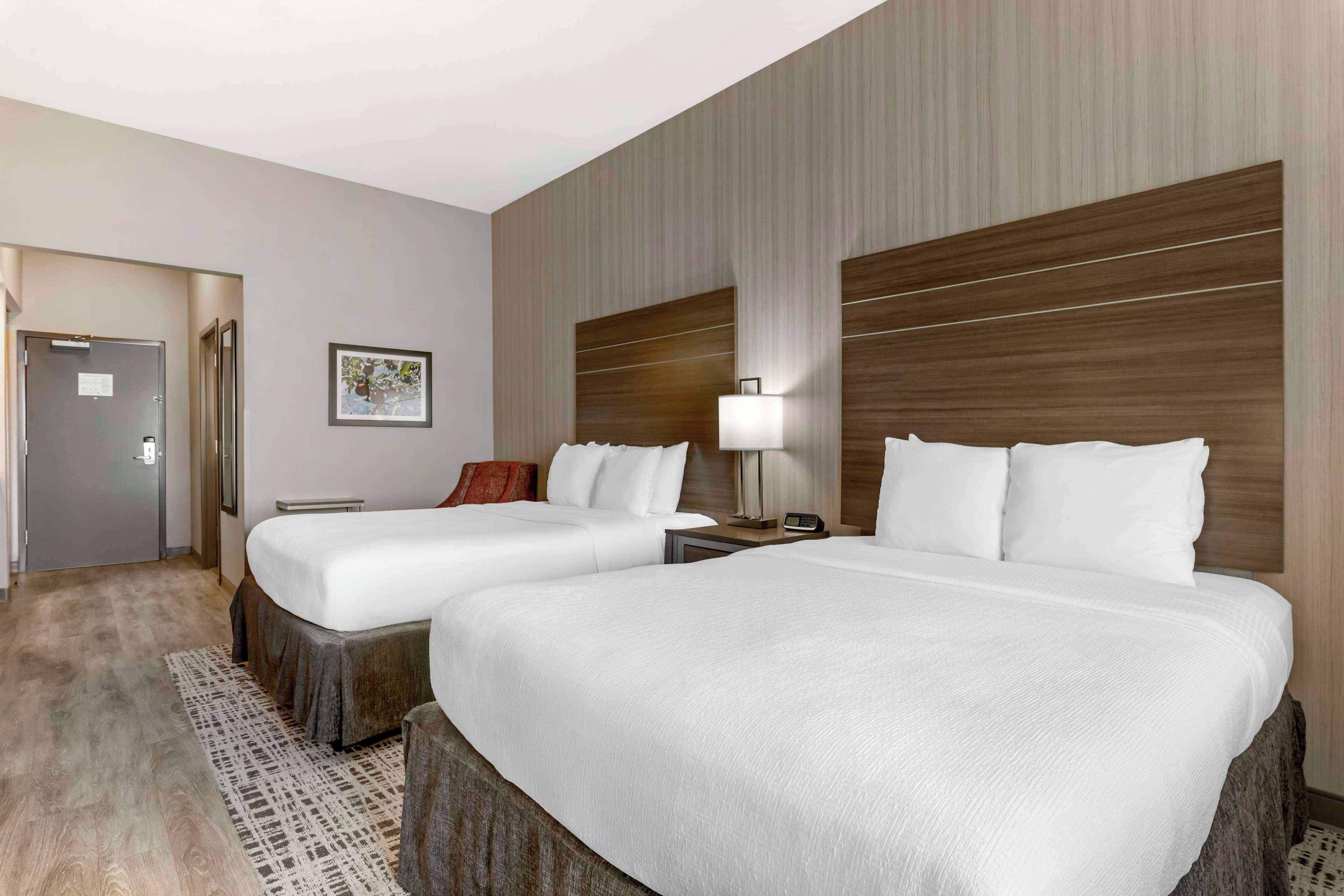 A hotel room with two beds, a wooden headboard, a bedside table with a lamp, and a framed picture on the wall.