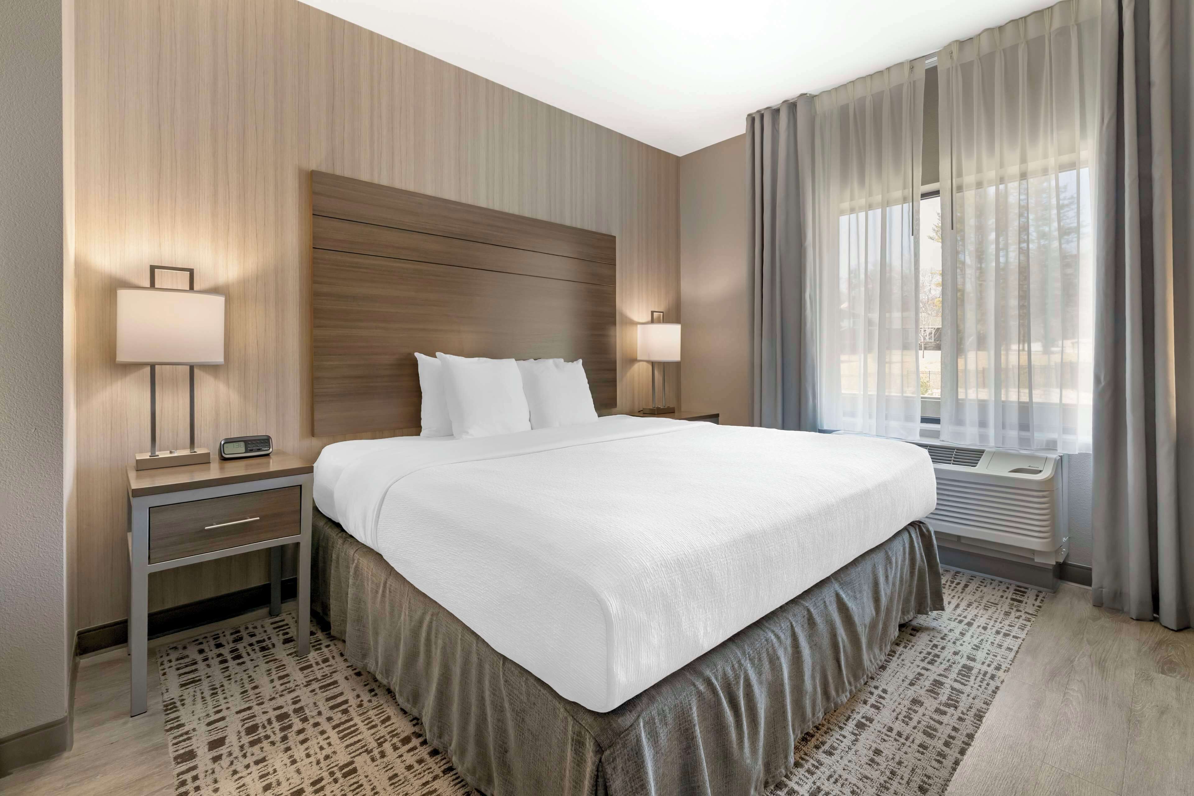 Quarto de hotel com uma cama grande, duas mesas de cabeceira com luminárias, uma cabeceira de madeira e uma janela ampla com cortinas.