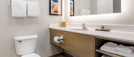 A modern bathroom with a toilet, sink, and mirror. There are two towels hanging on the wall and a shelf with folded towels.
