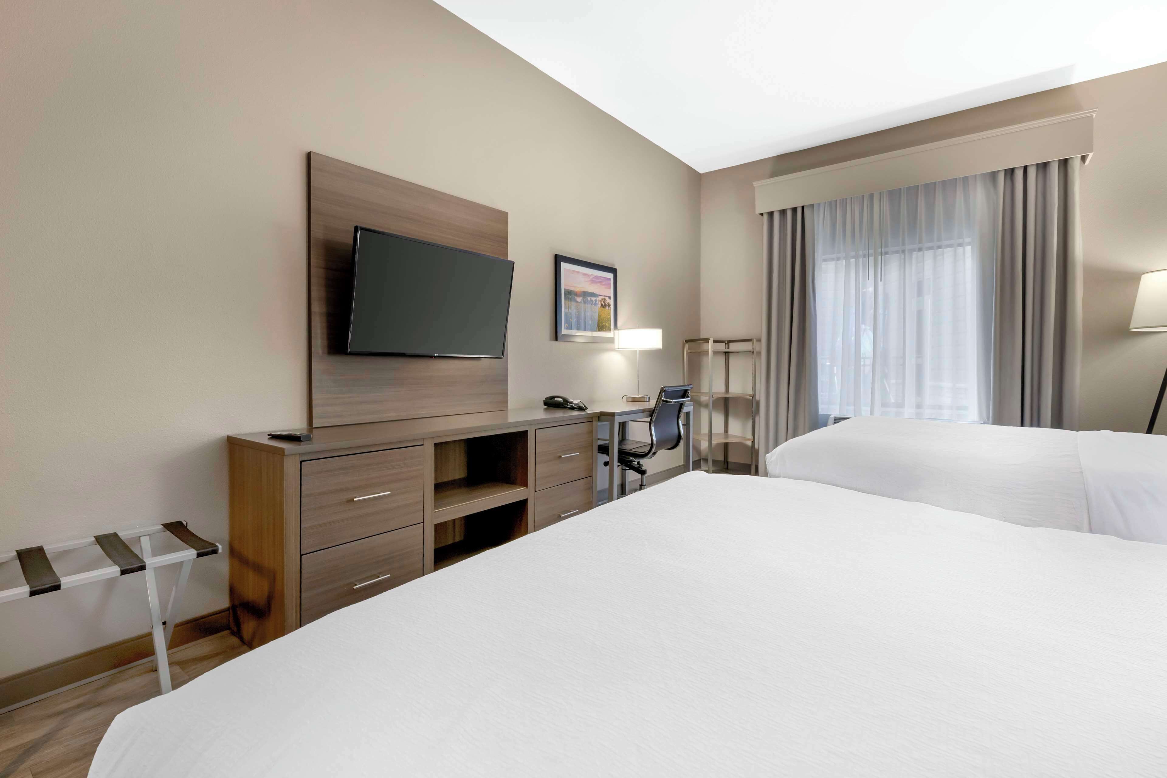 A hotel room with a large bed, a desk with a chair, a flat-screen TV mounted on the wall, a desk lamp, and a window with curtains.
