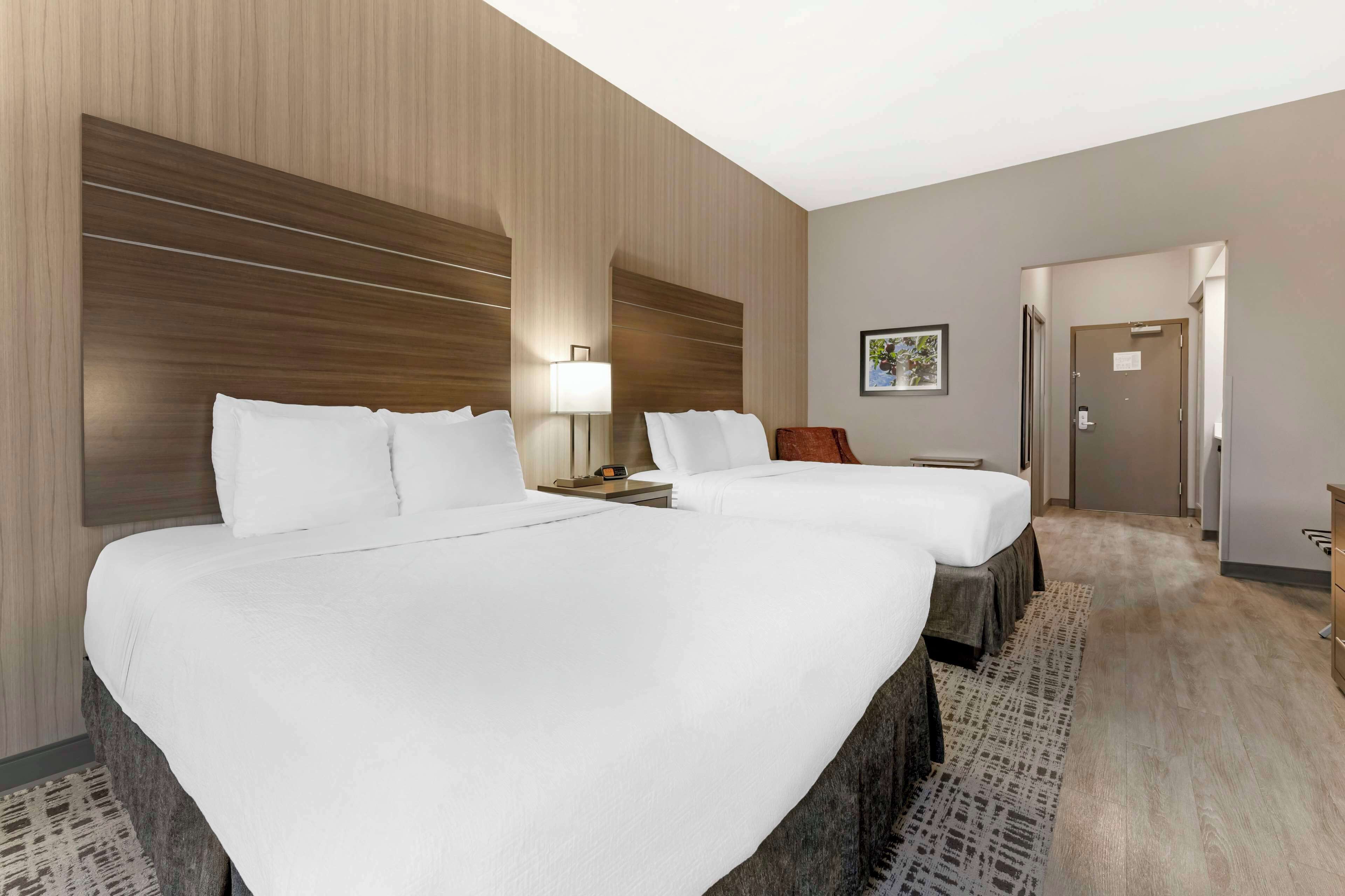 Una habitación de hotel con dos camas, una mesita de noche, una lámpara, un cuadro en la pared y una puerta que da a otra habitación.