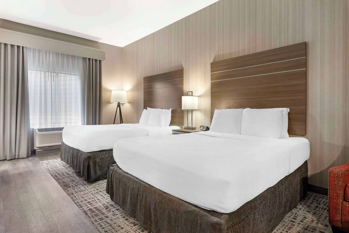A hotel room with two beds, a large window with curtains, a standing lamp, and a red chair.
