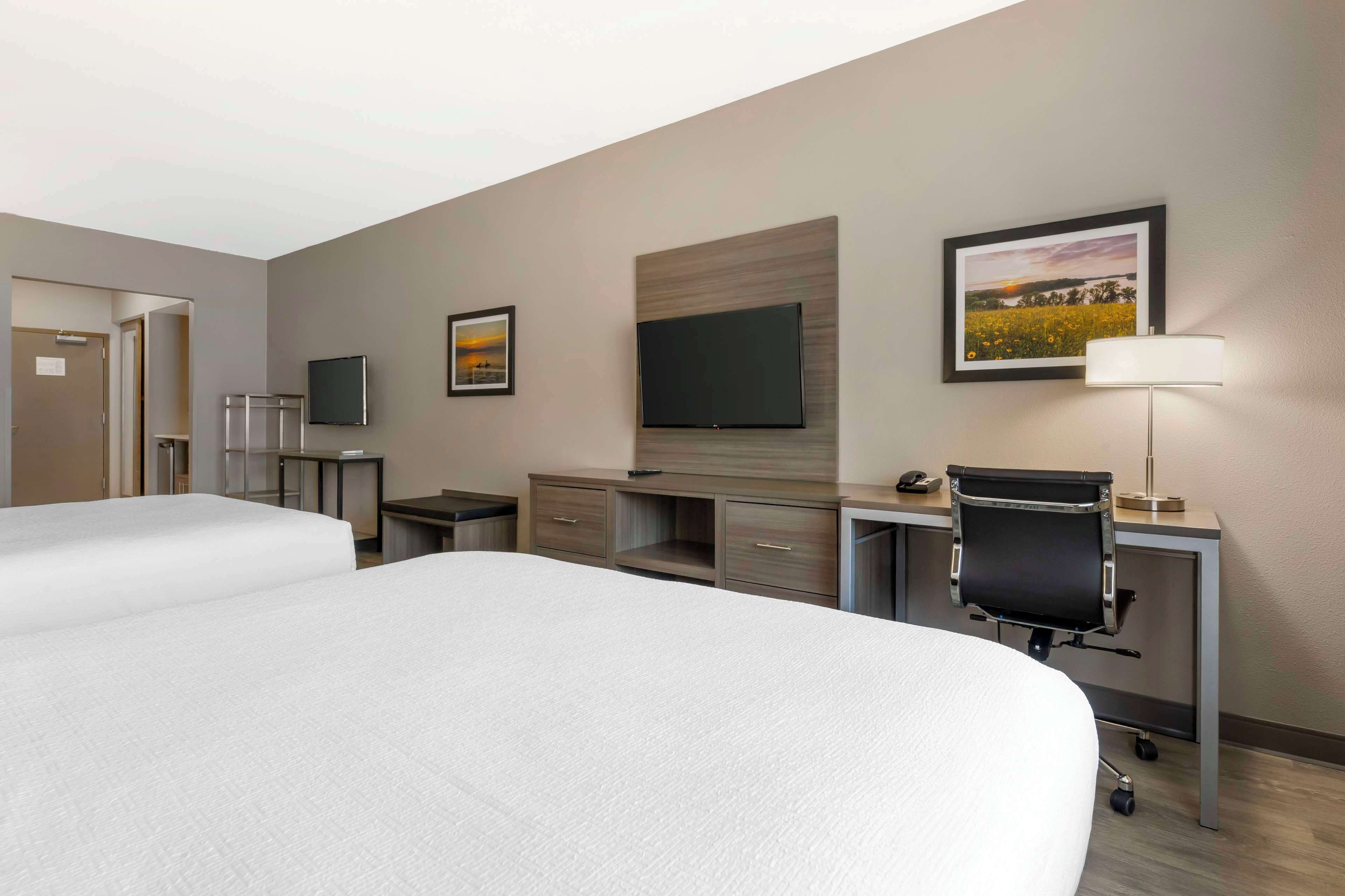 A hotel room with two beds, a desk with a chair, a flat-screen TV, a lamp, and framed pictures on the wall.