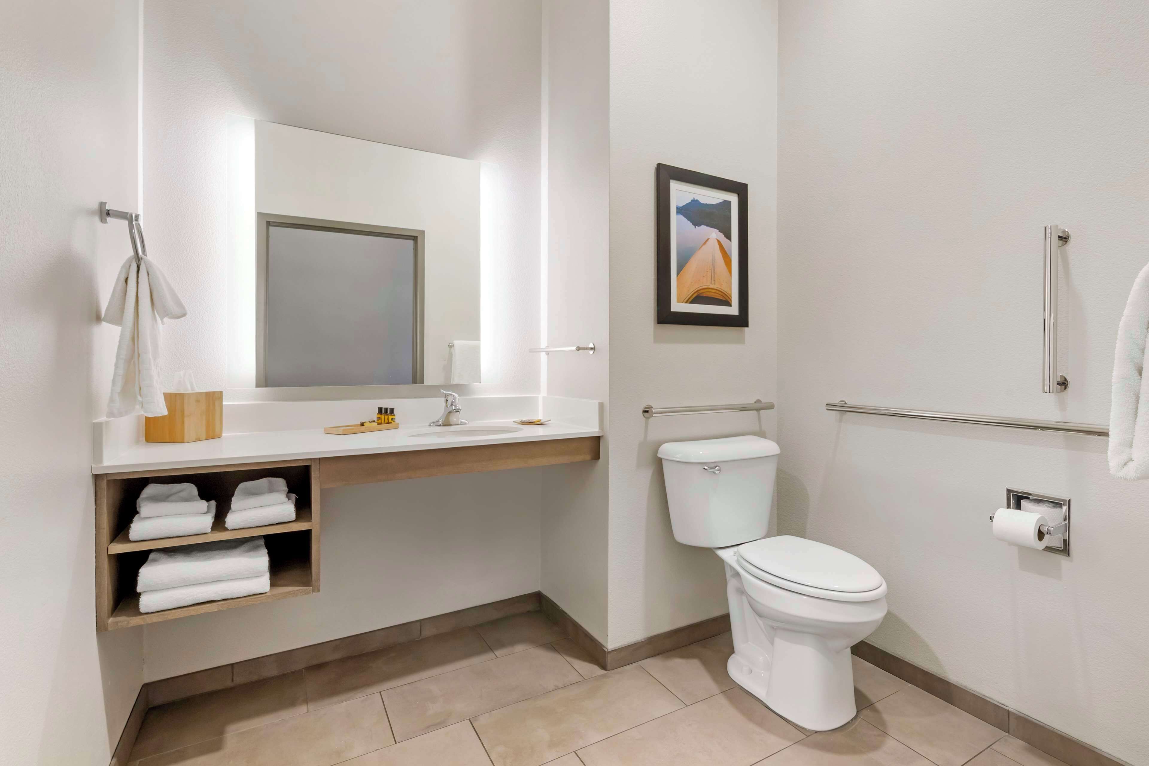 Une salle de bain moderne avec des toilettes, un lavabo et un miroir. On y trouve des étagères avec des serviettes et un tableau encadré accroché au mur.
