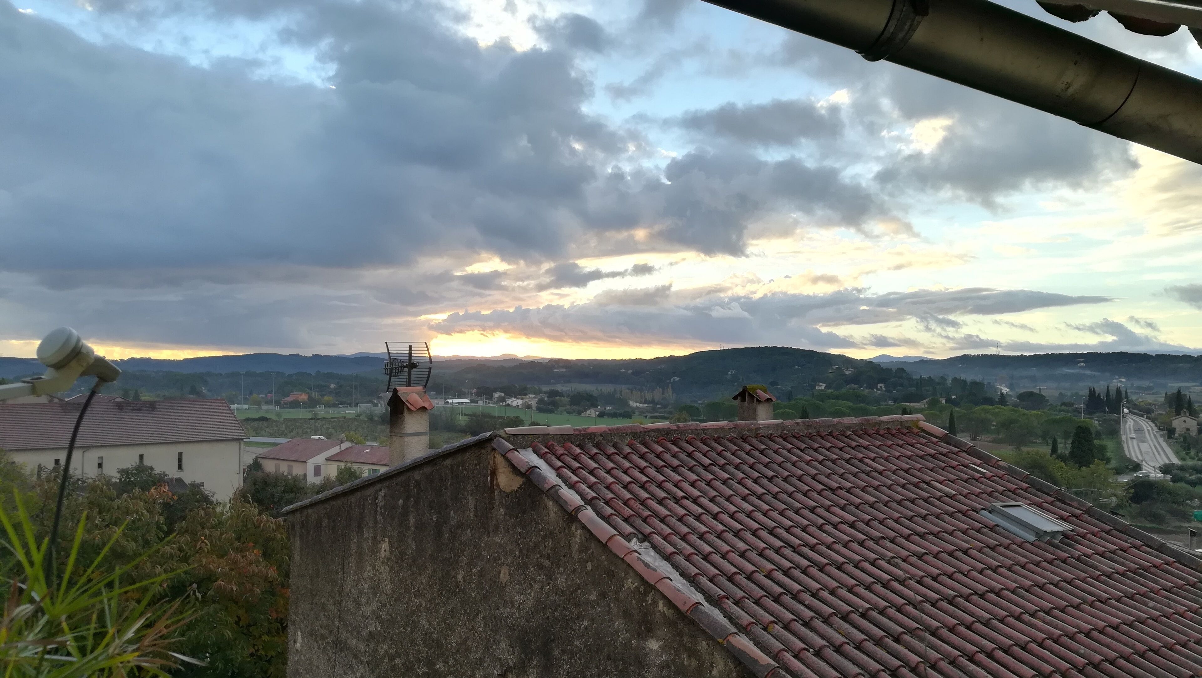 A view of a residential area with terracotta-tiled roofs, a satellite dish, and a distant sunset over rolling hills.