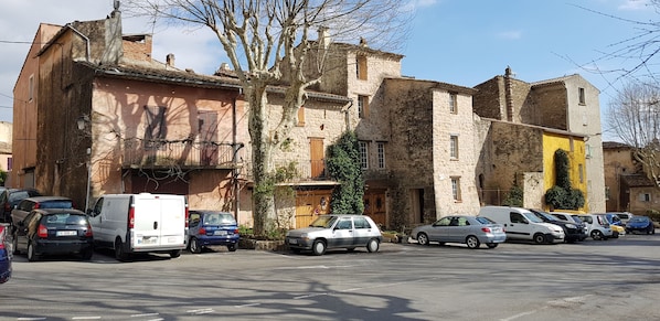 Una fila de coches aparcados a lo largo de una calle con edificios de piedra, algunos con balcones y vegetación.